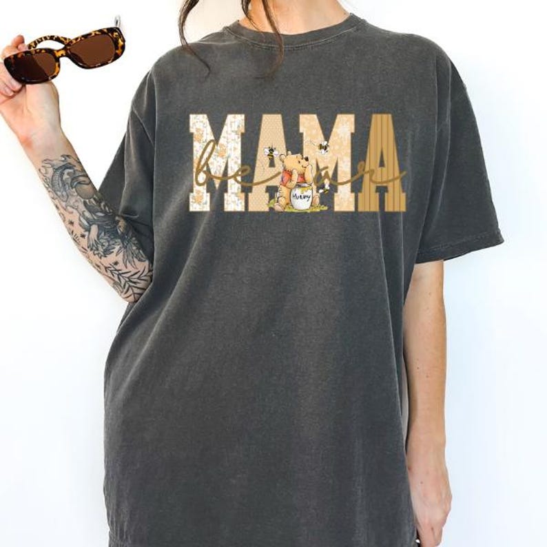May include: Dark gray t-shirt with the word "MAMA" in a block letter style, with "bee" in cursive script. The design includes a cartoon bear holding a honey pot, with bees. The shirt is a comfortable, casual style.