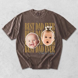 Może przedstawiać: Brązowa koszulka z napisem "BEST DAD EVER" w kolorze złotym. Koszulka przedstawia dwie twarze dziecka oraz imiona "Noah & Emily" w ozdobnej czcionce. Koszulka ma wygląd vintage.
