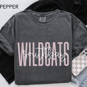 May include: A dark gray long-sleeve shirt with the text "WILDCATS southbay" in a light pink color. The word "PEPPER" is in the top left corner. A phone case with a checkered pattern is visible.