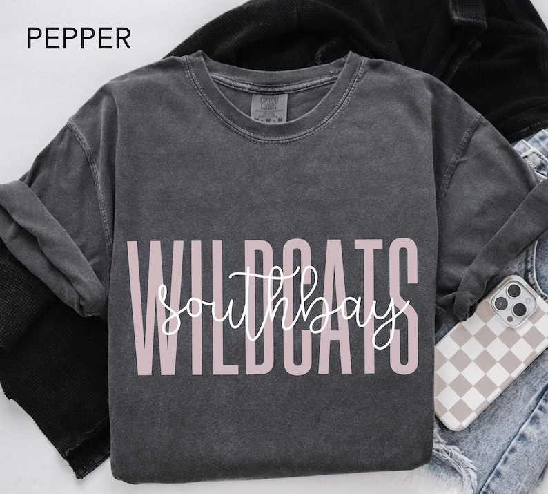 May include: A dark gray long-sleeve shirt with the text "WILDCATS southbay" in a light pink color. The word "PEPPER" is in the top left corner. A phone case with a checkered pattern is visible.