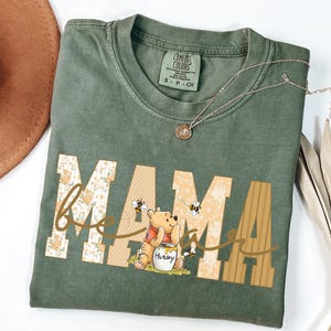 May include: Green Comfort Colors t-shirt with the word "MAMA" in a floral and tan design, with "bee" in cursive and a Winnie the Pooh graphic. A gold necklace is draped across the shirt.