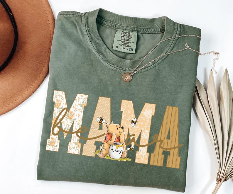 May include: Green Comfort Colors t-shirt with the word "MAMA" in a floral and tan design, with "bee" in cursive and a Winnie the Pooh graphic. A gold necklace is draped across the shirt.