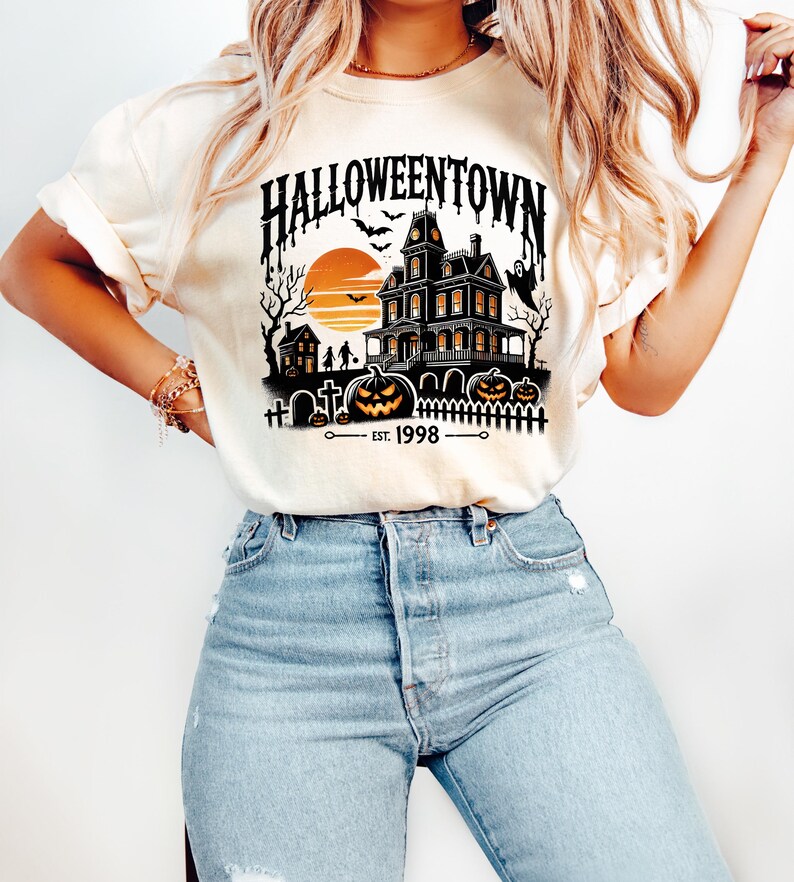 K&ouml;nnte beinhalten: Ein wei&szlig;es T-Shirt mit einem schwarzen Motiv eines Spukhauses, K&uuml;rbissen und Flederm&auml;usen. Der Text "HALLOWEENTOWN EST. 1998" ist auf dem Shirt aufgedruckt.