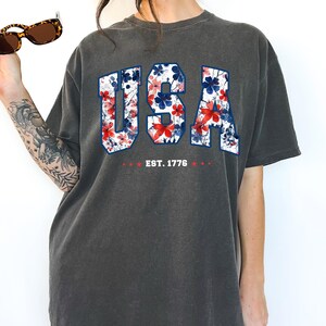 May include: Dark gray t-shirt with the word "USA" in a floral pattern of red and blue flowers. The text "EST. 1776" is below the word "USA". The shirt has short sleeves and a crew neck.