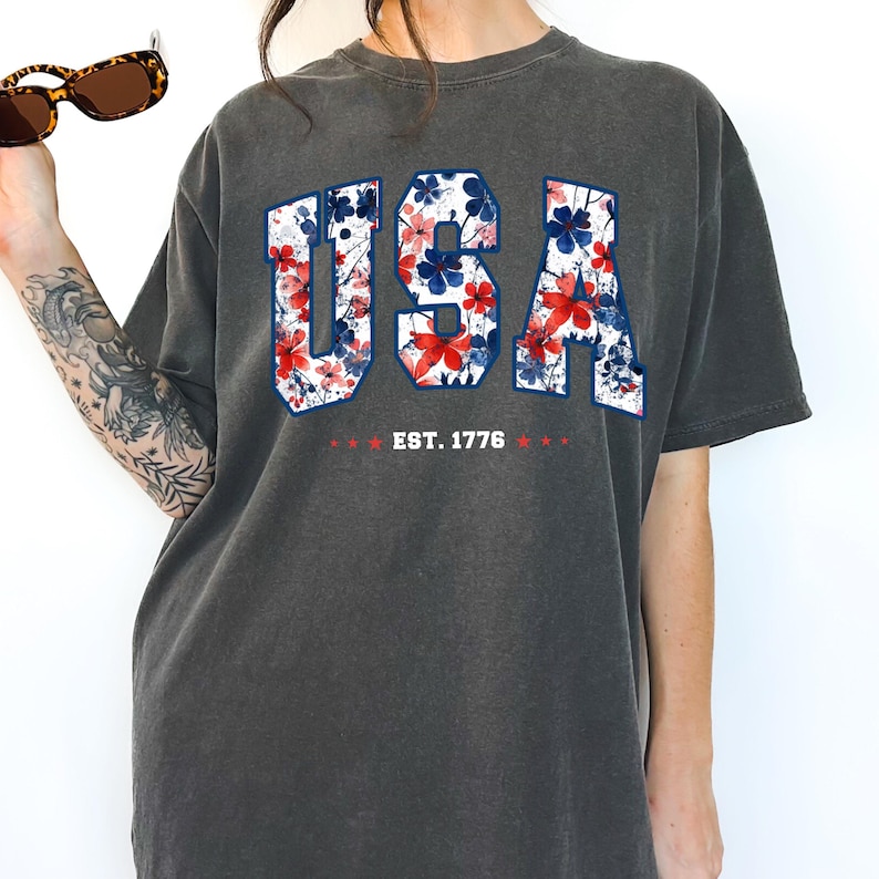 May include: Dark gray t-shirt with the word "USA" in a floral pattern of red and blue flowers. The text "EST. 1776" is below the word "USA". The shirt has short sleeves and a crew neck.