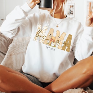 Op de afbeelding: Witte sweatshirt met het woord "Mama" in een beige en bruin ontwerp, met een cartoonbeer en de tekst "EST. 2026". De sweatshirt heeft lange mouwen en een ronde hals.