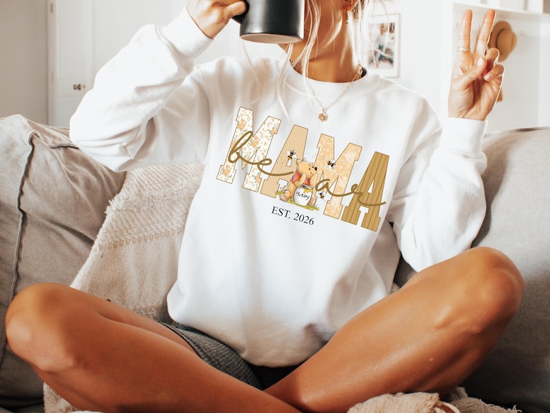 Op de afbeelding: Witte sweatshirt met het woord "Mama" in een beige en bruin ontwerp, met een cartoonbeer en de tekst "EST. 2026". De sweatshirt heeft lange mouwen en een ronde hals.