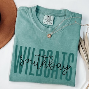May include: A teal-colored t-shirt with the words "WILDCATS southbay" printed in dark green and black script. The shirt has a crew neck and short sleeves. A silver necklace with a small pendant rests on the shirt.