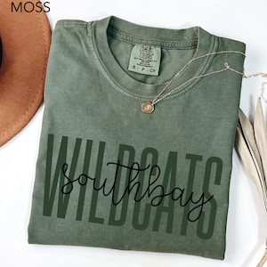 May include: A moss green t-shirt with the words "WILDCATS southbay" printed in black. The shirt has a crew neck and short sleeves. A gold necklace with a pendant is draped across the shirt.