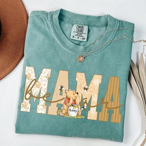 May include: A teal Comfort Colors t-shirt with the word "MAMA" in a floral and dotted design, with "be" in cursive and a Winnie the Pooh graphic. A silver necklace with a pendant is on the shirt.