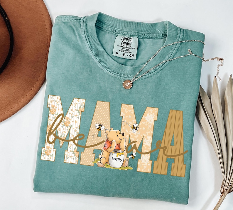 May include: A teal Comfort Colors t-shirt with the word "MAMA" in a floral and dotted design, with "be" in cursive and a Winnie the Pooh graphic. A silver necklace with a pendant is on the shirt.