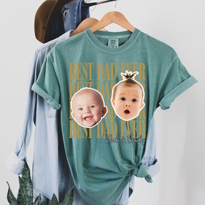 Może przedstawiać: Turkusowa koszulka z napisem "BEST DAD EVER" w złotym kolorze. Dwie twarze dziecka i imiona "Noah & Emily" są nadrukowane. Koszulka wisi na drewnianym wieszaku.