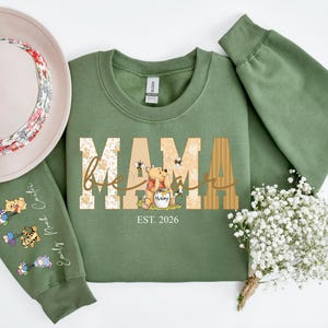Op de afbeelding: Een olijfgroene sweatshirt met het woord "MAMA" in een decoratief lettertype, met een Winnie de Poeh-afbeelding. De mouw heeft een aangepaste naam en een karakterontwerp. Een bloemenhoed staat op de achtergrond.