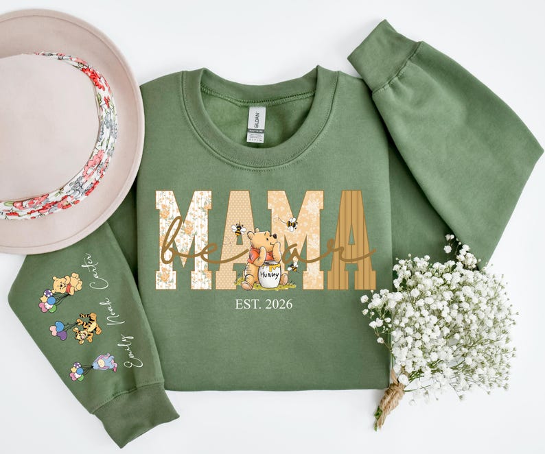 Op de afbeelding: Een olijfgroene sweatshirt met het woord "MAMA" in een decoratief lettertype, met een Winnie de Poeh-afbeelding. De mouw heeft een aangepaste naam en een karakterontwerp. Een bloemenhoed staat op de achtergrond.