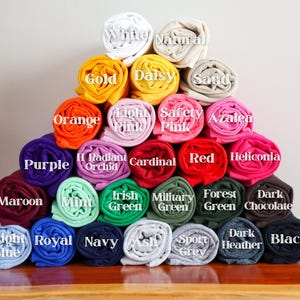 Op de afbeelding: Een piramide van opgerolde t-shirts in diverse kleuren, waaronder wit, goud, oranje, paars, bordeauxrood, lichtblauw, koningsblauw, marineblauw, asgrijs, sportgrijs, donkergrijs en zwart. Elke rol toont de kleurnaam.