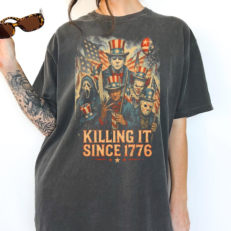 以下が含まれることがあります： ダークグレーのTシャツ。愛国的な衣装を着たホラー映画のキャラクターのグラフィックが特徴で、背景にはアメリカの国旗があります。「KILLING IT SINCE 1776」のテキストが下に印刷されています。