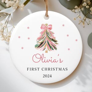 May include: White ceramic Christmas ornament with a pink and green watercolor Christmas tree, pink bow, and gold glitter. The ornament reads "Olivia's First Christmas 2024".