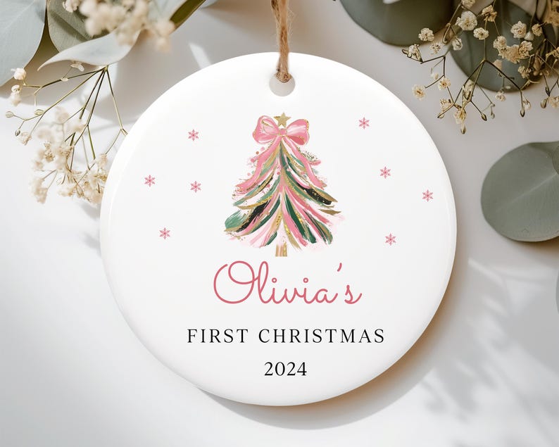 May include: White ceramic Christmas ornament with a pink and green watercolor Christmas tree, pink bow, and gold glitter. The ornament reads "Olivia's First Christmas 2024".