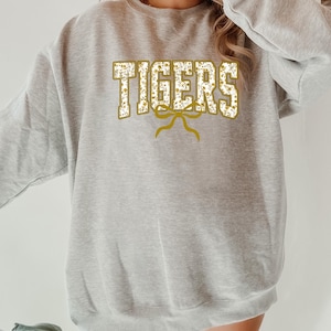 K&ouml;nnte beinhalten: Ein hellgraues Sweatshirt mit dem Wort "TIGERS" in goldener, floral gef&uuml;llter Schrift. Unter dem Wort befindet sich ein goldenes Schleifendesign. Das Sweatshirt ist ein l&auml;ssiger Stil.