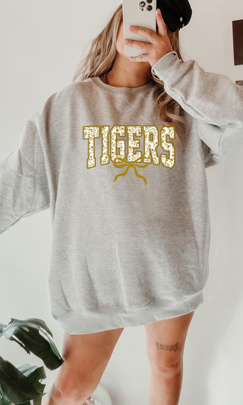 K&ouml;nnte beinhalten: Ein hellgraues Sweatshirt mit dem Wort "TIGERS" in goldener, floral gef&uuml;llter Schrift. Unter dem Wort befindet sich ein goldenes Schleifendesign. Das Sweatshirt ist ein l&auml;ssiger Stil.