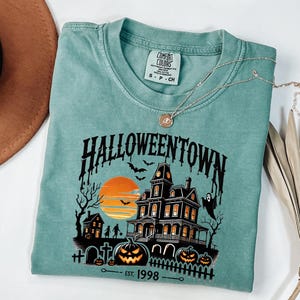 K&ouml;nnte beinhalten: Ein blaues T-Shirt mit einem Grafikdruck eines Spukhauses mit einem Sonnenuntergang im Hintergrund. Der Text "HALLOWEENTOWN - EST. 1998" ist unter dem Grafikdruck aufgedruckt.
