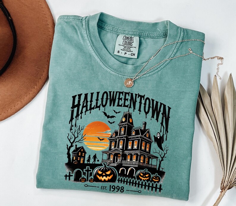 K&ouml;nnte beinhalten: Ein blaues T-Shirt mit einem Grafikdruck eines Spukhauses mit einem Sonnenuntergang im Hintergrund. Der Text "HALLOWEENTOWN - EST. 1998" ist unter dem Grafikdruck aufgedruckt.