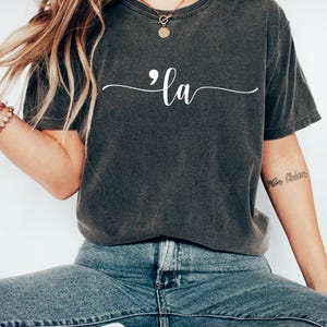 May include: A dark gray t-shirt with a white semicolon and the word "la" written in cursive.