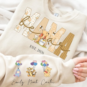 Op de afbeelding: Cr&egrave;mekleurige sweatshirt met het woord "MAMA" in bloemen- en houtnerfdesign, met Winnie de Poeh en bijen. De mouw toont stripfiguren met ballonnen en de namen "Emily Noah Carter". De tekst "EST. 2026" is ook zichtbaar.