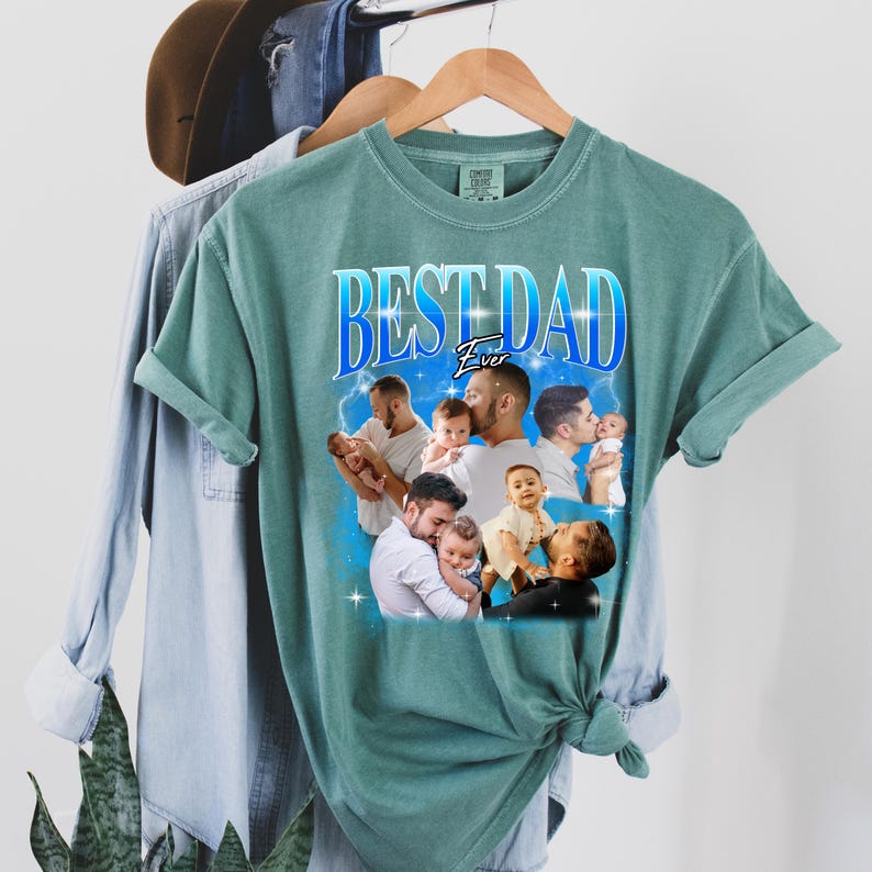 May include: A teal-colored t-shirt with the text "BEST DAD Ever" in a blue and white design. The shirt features a collage of photos of a father with his child. The shirt is hanging on a wooden hanger with other clothing items in the background.