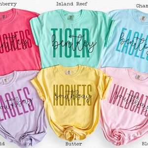 May include: Six colorful t-shirts in shades of pink, green, blue, purple, yellow, and pink. Each shirt features a team name and the word "Bentley" or "Southbay" in a contrasting color. The shirts are folded.