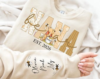 May include: Cream-colored sweatshirt with the word "NANA" in a floral and wood-grain design, featuring Winnie the Pooh and bees. The text "EST. 2026" is below the graphic. The sleeve displays names "Olivia, Noah, Lily" with bee illustrations.