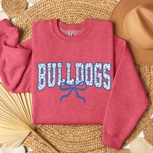 K&ouml;nnte beinhalten: Ein rotes Sweatshirt mit dem Wort "BULLDOGS" in wei&szlig;en Buchstaben mit blauem Blumenmuster. Unter dem Wort befindet sich eine blaue Schleife. Das Sweatshirt wird auf einer gewebten Oberfl&auml;che mit einem braunen Hut pr&auml;sentiert.