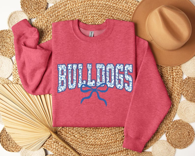 K&ouml;nnte beinhalten: Ein rotes Sweatshirt mit dem Wort "BULLDOGS" in wei&szlig;en Buchstaben mit blauem Blumenmuster. Unter dem Wort befindet sich eine blaue Schleife. Das Sweatshirt wird auf einer gewebten Oberfl&auml;che mit einem braunen Hut pr&auml;sentiert.