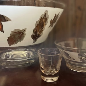 May include: A large glass punch bowl, a smaller glass bowl, and a small glass cup. Each piece features a white band decorated with metallic gold leaf patterns. The glass is clear, and the metallic gold leaves are arranged in a repeating design.
