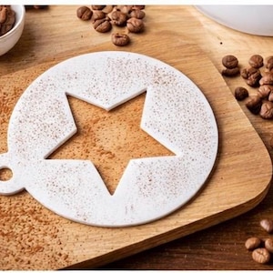 May include: A white coffee stencil with a star cutout, dusted with brown powder, rests on a wooden cutting board. Coffee beans and a small bowl of cocoa powder surround the stencil, suggesting its use for decorative coffee art.