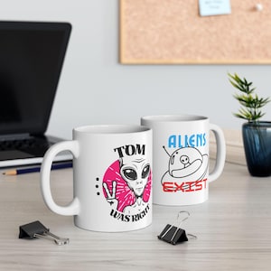 May include: Two white ceramic mugs with alien-themed designs. One mug features an alien illustration with the text "TOM WAS RIGHT". The other mug has a UFO illustration with the text "ALIENS EXIST".