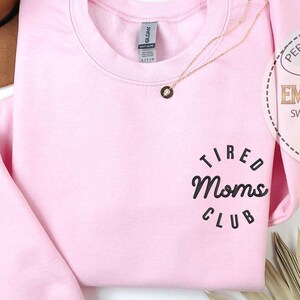 May include: Pink sweatshirt with the text "Tired Moms Club" in a black circle on the front. The sweatshirt is a crew neck style and has a relaxed fit.