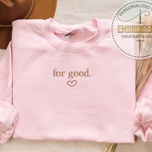 May include: A pink sweatshirt with the text "for good." embroidered in brown on the front. A small heart is embroidered below the text.