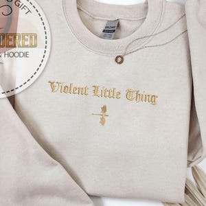 May include: A beige sweatshirt with the embroidered text "Violent Little Thing" in gold. A gold necklace with a circular pendant rests on the sweatshirt. The image also includes a decorative fan and a personalized gift label.