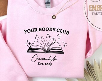 May include: A light pink sweatshirt with the text "YOUR BOOKS CLUB" above an open book graphic. Below the graphic is "One more chapter EST. 2025". A gold necklace and a decorative leaf are also visible.