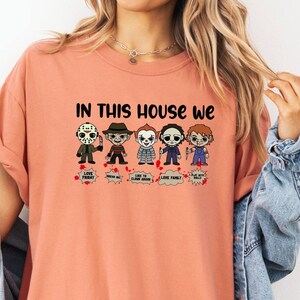 May include: A peach colored t-shirt with the text "In this house we" and a row of five cartoon horror characters. The characters are wearing different outfits and have different facial expressions.