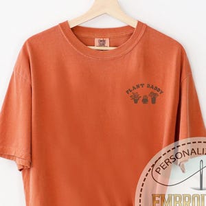 May include: A rust-colored t-shirt with the text "Plant Daddy" embroidered on the front. The shirt is on a hanger against a white wall. The text "Personalized Gift" and "Embroidered Comfort Colors" is visible in a stitched circle.