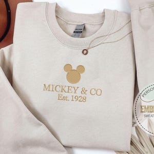 May include: Beige sweatshirt with embroidered Mickey Mouse logo and "Mickey & Co Est. 1928" text. A gold necklace and a brown hat are also visible. The sweatshirt is part of a personalized gift set.