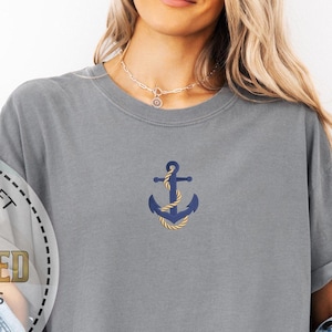 May include: A gray t-shirt with a navy blue anchor embroidered on the chest. The anchor is detailed with gold rope accents. The shirt has a relaxed fit and is part of the Comfort Colors collection. The image also includes the text "Personalized Gift" and "Embroidered".