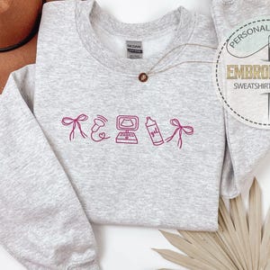 May include: A light gray sweatshirt with embroidered pink graphics and text. The design includes a bow, a heart, a computer, a bottle, and a bow. A brown hat and a necklace are also visible. The sweatshirt has the text "Personalized Gift" and "Embroidered Sweatshirt & Hoodie".