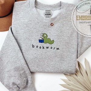 May include: A heather gray sweatshirt with a crew neck and long sleeves. The front features a green cartoon bookworm wearing glasses and holding a book, with the word "bookworm" below. A brown hat is visible in the background.
