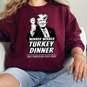 May include: Burgundy sweatshirt with a graphic of a man holding a fist up, with the text "WINNER WINNER TURKEY DINNER MAKE THANKSGIVING GREAT AGAIN!". The sweatshirt is a solid color and has long sleeves.