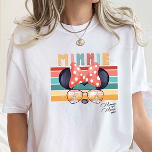 Op de afbeelding: Wit T-shirt met een retro-stijl grafische afbeelding van Minnie Mouse met een zonnebril en een rode strik. De afbeelding bevat de tekst "Minnie" en een regenboogstrepenpatroon.