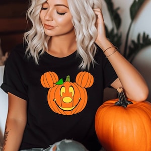 May include: A black t-shirt with a pumpkin-shaped Mickey Mouse head graphic. The pumpkin has a smiling face with a black nose and green stem. The shirt is being worn by a person with blonde hair.