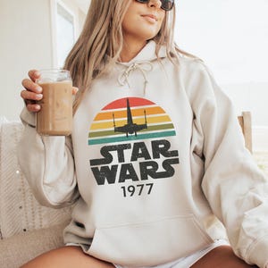 May include: Cream-colored Star Wars hoodie with a retro sunset graphic. The design features a silhouette of a spaceship and the text "STAR WARS 1977". The hoodie is worn by a person wearing sunglasses and holding a glass of iced coffee.